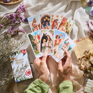May include: A deck of Magna Bloom Tarot cards with floral and botanical illustrations. The cards are held in hands, fanned out to display the artwork. The box features the title "Magna Bloom" with floral designs.