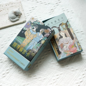May include: A blue box with a gold border and the text "Quest Enlightenment Cards" on the front. The box contains a deck of cards with illustrations of people and flowers. The back of the box has the text "Quest Enlightenment Cards" in Chinese characters.