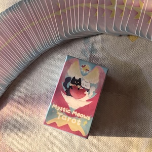 K&ouml;nnte beinhalten: Ein Kartenspiel Mystic Meows Tarot mit einer Schachtel mit einer skurrilen Illustration von zwei Katzen. Die Schachtel hat einen rosa und blauen Hintergrund mit den Worten "Mystic Meows Tarot". Die Karten haben rosa und gelbe Akzente.