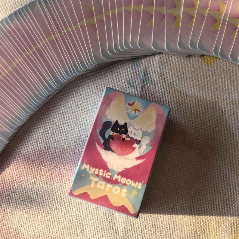 K&ouml;nnte beinhalten: Ein Kartenspiel Mystic Meows Tarot mit einer Schachtel mit einer skurrilen Illustration von zwei Katzen. Die Schachtel hat einen rosa und blauen Hintergrund mit den Worten "Mystic Meows Tarot". Die Karten haben rosa und gelbe Akzente.