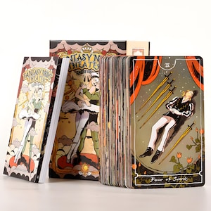 May include: A tarot card deck with a fantasy theme. The deck includes a box, a guidebook, and a deck of cards. The cards feature illustrations of ballet dancers and other theatrical figures. The "Four of Swords" card is visible, with a figure lying down.