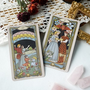 May include: Two tarot cards, one with the number 10 and the other with the number 2. The card with the number 10 depicts a family of four with a dark blue sky and a rainbow of seashells above them. The card with the number 2 depicts a couple in medieval clothing holding cups, with a floral background.