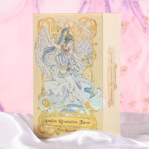 May include: A beige tarot card box featuring an illustration of a winged figure in flowing white and blue robes, holding a golden chalice. The box is labeled "Avalon Revelation Tarot" and "Taru Hanasaki".