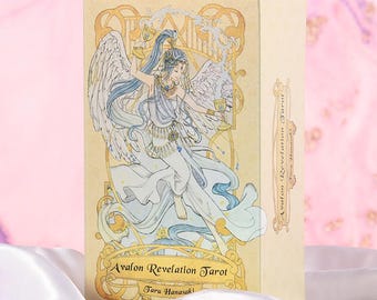 May include: A beige tarot card box featuring an illustration of a winged figure in flowing white and blue robes, holding a golden chalice. The box is labeled "Avalon Revelation Tarot" and "Taru Hanasaki".