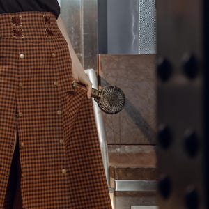 May include: A brown and black plaid skirt with a lace-up detail at the waist and a front slit. The skirt is worn by a person holding a small, handheld fan.