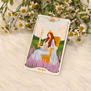 May include: A tarot card featuring a woman with long red hair wearing a crown and a blue dress, holding a sword. She is seated on a yellow chair, with a background of green hills and lavender. The card has a white border with a gold trim.