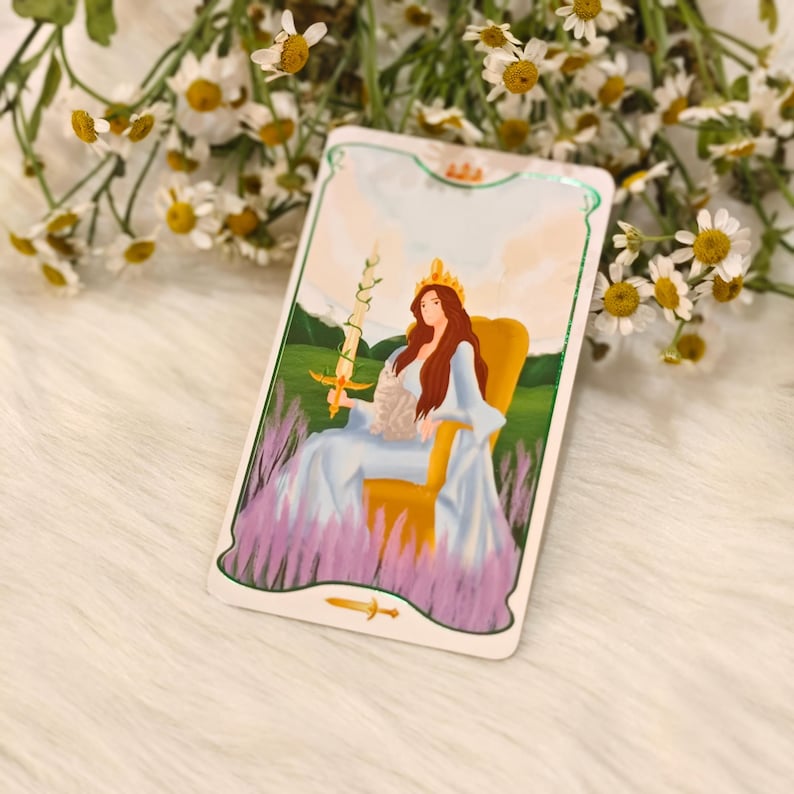 May include: A tarot card featuring a woman with long red hair wearing a crown and a blue dress, holding a sword. She is seated on a yellow chair, with a background of green hills and lavender. The card has a white border with a gold trim.