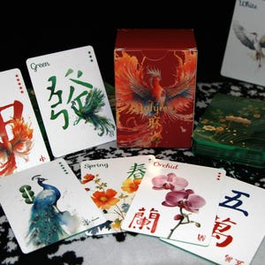 Pu&ograve; includere: Un set di Mahjong con una scatola rossa con un design a fenice e carte con illustrazioni floreali e di uccelli. Le carte presentano caratteri cinesi e parole inglesi come "Green", "Spring" e "Orchid".