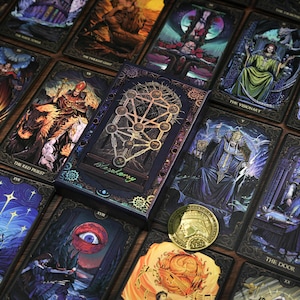 May include: A set of tarot cards with detailed illustrations and a matching box. The cards display artwork in blue, purple, gold, and brown tones. The box is dark blue with a gold design and the word "Blasphemy". A gold coin is also present.