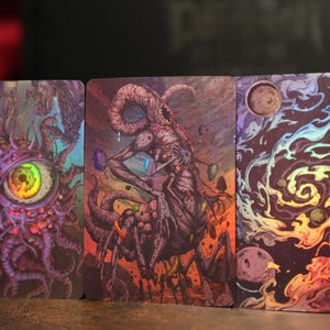 May include: Three rectangular trading cards featuring vibrant, detailed illustrations. The left card shows a large, central eye surrounded by purple tentacles. The middle card depicts a multi-limbed creature. The right card displays a colorful galaxy.