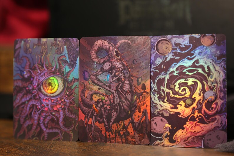 May include: Three rectangular trading cards featuring vibrant, detailed illustrations. The left card shows a large, central eye surrounded by purple tentacles. The middle card depicts a multi-limbed creature. The right card displays a colorful galaxy.