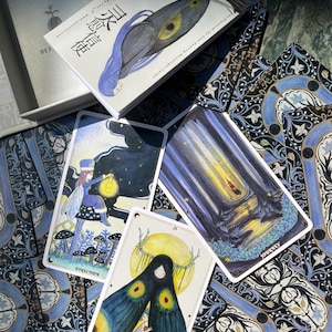May include: A deck of oracle cards with a box featuring a moth illustration. The cards have illustrations of fairies, forests, and a moth-like figure. The cards are on a patterned blue and white surface.