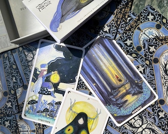 Könnte beinhalten: Ein Orakelkarten-Deck mit einer Schachtel mit einer Mottenillustration. Die Karten zeigen Illustrationen von Feen, Wäldern und einer mottenartigen Figur. Die Karten liegen auf einer gemusterten blau-weißen Oberfläche.