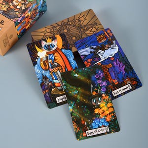 May include: A tarot card deck with colorful illustrations. The cards feature whimsical artwork, including a cat king, a whale, and other fantasy elements. The deck is displayed with a wooden box, and the cards are arranged to showcase the unique designs.