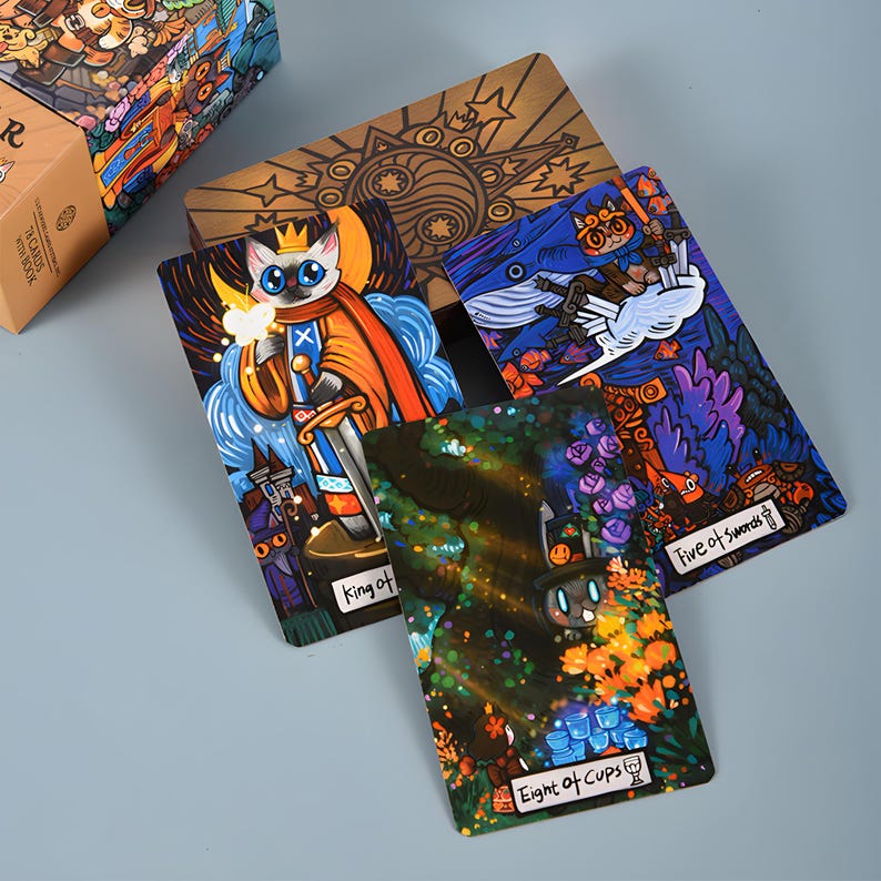 May include: A tarot card deck with colorful illustrations. The cards feature whimsical artwork, including a cat king, a whale, and other fantasy elements. The deck is displayed with a wooden box, and the cards are arranged to showcase the unique designs.