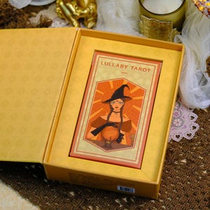 K&ouml;nnte beinhalten: Ein Tarotkartenspiel mit dem Titel "Lullaby Tarot" in einer gelben Box. Die Karte zeigt eine Illustration einer Hexe mit schwarzem Hut, Z&ouml;pfen und einer Kristallkugel. Die Box hat ein gemustertes Innenleben.