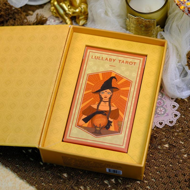 K&ouml;nnte beinhalten: Ein Tarotkartenspiel mit dem Titel "Lullaby Tarot" in einer gelben Box. Die Karte zeigt eine Illustration einer Hexe mit schwarzem Hut, Z&ouml;pfen und einer Kristallkugel. Die Box hat ein gemustertes Innenleben.