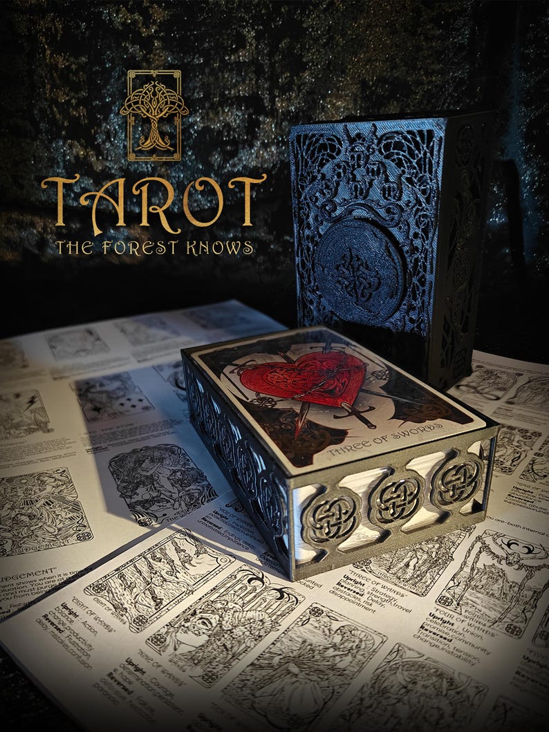 May include: A tarot card box with a decorative design, featuring the text "THREE OF SWORDS" on the top card. The box is placed on a surface covered with tarot card illustrations. The image also includes a matching box and the text "TAROT THE FOREST KNOWS".