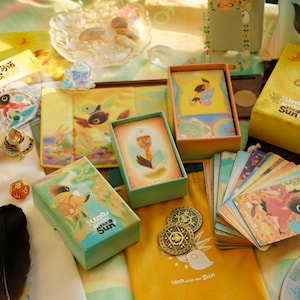 May include: An assortment of illustrated tarot cards, boxes, and accessories, including dice, feathers, and coins. The products feature a bird-like character and the text "Uno the Sun" in a whimsical, colorful design. The overall theme is a spiritual or fantasy-related product.