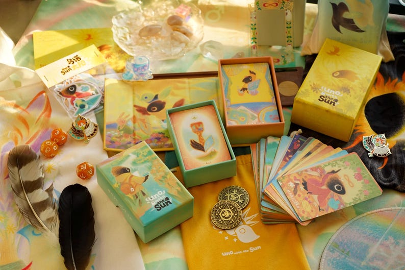 May include: An assortment of illustrated tarot cards, boxes, and accessories, including dice, feathers, and coins. The products feature a bird-like character and the text "Uno the Sun" in a whimsical, colorful design. The overall theme is a spiritual or fantasy-related product.