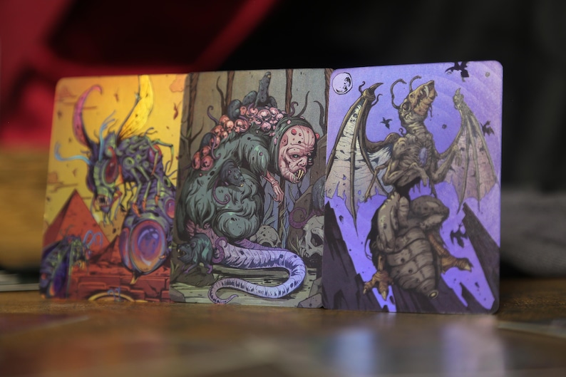 May include: Three rectangular trading cards featuring detailed, colorful illustrations of monstrous creatures. The left card shows a yellow-toned insectoid beast, the center card depicts a green, multi-eyed creature, and the right card shows a winged, skeletal monster on a purple background.