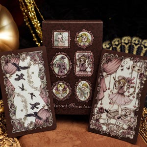 Op de afbeelding: Een Vacant House tarotkaartenset met een doosje en twee kaarten voorzien van bruine lijsten, roze gordijnen en illustraties van figuren en kraaien. De rechterkaart is gelabeld The Fool en toont een figuur in een roze jurk met vlinders.