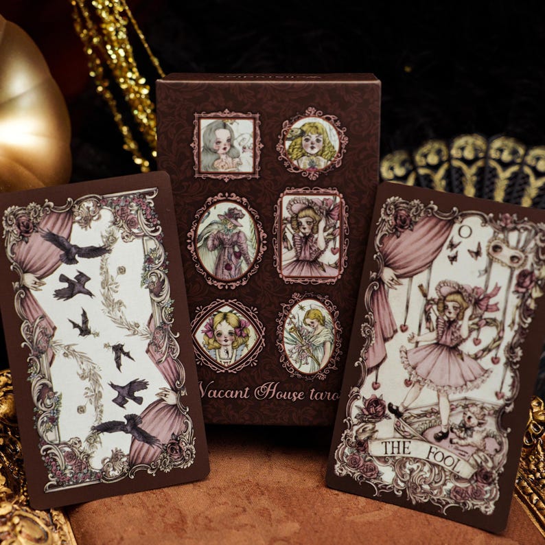 May include: A Vacant House tarot deck box and two cards featuring ornate brown frames, pink curtains, and illustrations of figures and crows. The right card is labeled The Fool, showing a character in a pink dress with butterflies and a small dog.