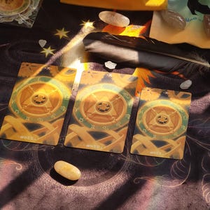 May include: Three tarot cards with a golden wheel design, the word "WHEEL" at the bottom, and a feather on a patterned surface. The cards are arranged with crystals and other decorative elements.