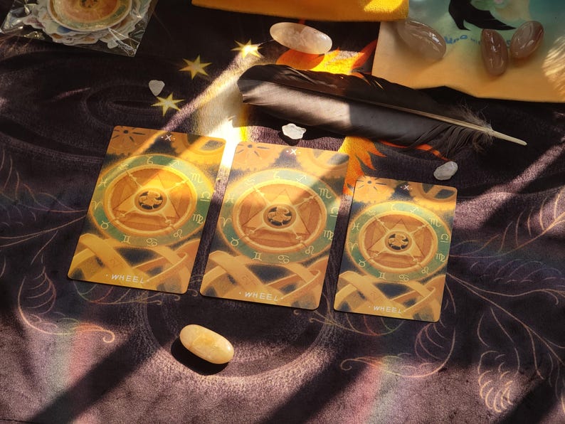 May include: Three tarot cards with a golden wheel design, the word "WHEEL" at the bottom, and a feather on a patterned surface. The cards are arranged with crystals and other decorative elements.