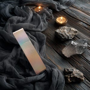 May include: A rectangular, iridescent metal bar with a smooth, reflective surface that shifts in colour from soft gold to light purple. The object is positioned diagonally against a dark, textured fabric backdrop with two lit candles and three raw stones.
