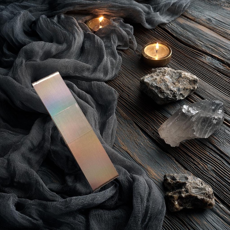 May include: A rectangular, iridescent metal bar with a smooth, reflective surface that shifts in colour from soft gold to light purple. The object is positioned diagonally against a dark, textured fabric backdrop with two lit candles and three raw stones.