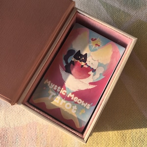 K&ouml;nnte beinhalten: Ein Tarotkartenspiel mit dem Titel "Mystic Meows Tarot" in einer rosa Schachtel. Das Kartendesign zeigt zwei Zeichentrickkatzen, eine schwarze und eine wei&szlig;e, auf einem rosa und blauen Hintergrund. Die Schachtel ist hellrosa mit wei&szlig;em Innenraum.