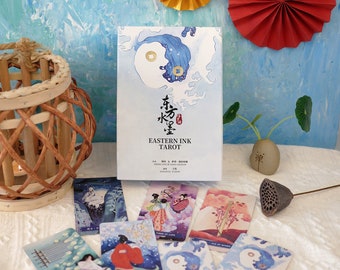 Pode incluir: Uma caixa de cartas de tarô Eastern Ink com um design azul e branco. A caixa apresenta um símbolo yin yang e o texto "Eastern Ink Tarot" em preto. As cartas estão espalhadas sobre uma superfície branca com um fundo azul.