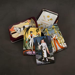May include: A tarot card deck with a burgundy metal tin. The tin features a woman in a white suit with the text "Fantasy Theatre". Several tarot cards are visible, each with unique illustrations and titles like "The Magician" and "The Hermit".