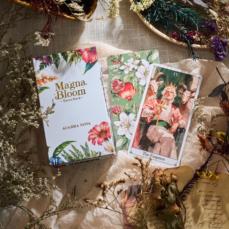 May include: A white tarot deck box with floral illustrations and the text "Magna Bloom Tarot Deck" and "ACABRA NOVA". Two tarot cards are visible, one with "The Emperor" and another with floral and pomegranate designs.