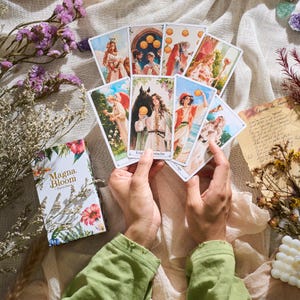 May include: A deck of Magna Bloom Tarot cards with floral and botanical illustrations. The cards are held in hands, displaying various images of figures and symbols. The box features the text "Magna Bloom - Tarot Deck".