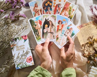May include: A deck of Magna Bloom Tarot cards with floral and botanical illustrations. The cards are held in hands, displaying various images of figures and symbols. The box features the text "Magna Bloom - Tarot Deck".