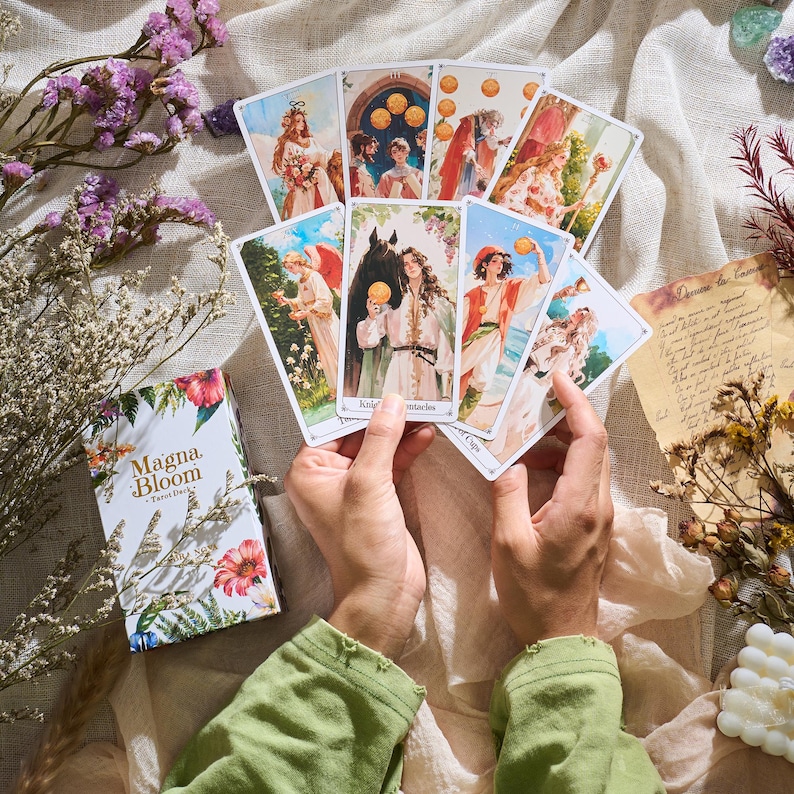 May include: A deck of Magna Bloom Tarot cards with floral and botanical illustrations. The cards are held in hands, displaying various images of figures and symbols. The box features the text "Magna Bloom - Tarot Deck".