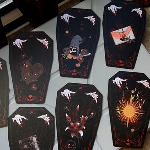 May include: A collection of coffin-shaped cards featuring dark illustrations with red accents. Each card displays a unique graphic, such as a heart, sun, moon, or letter, and includes pale hands with red fingernails at the top, appearing to manipulate strings.