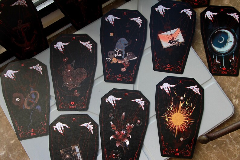 May include: A collection of coffin-shaped cards featuring dark illustrations with red accents. Each card displays a unique graphic, such as a heart, sun, moon, or letter, and includes pale hands with red fingernails at the top, appearing to manipulate strings.