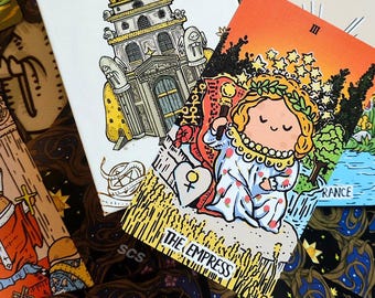 May include: A tarot card with an orange background and a cartoon illustration of a woman wearing a crown of stars. The card is titled "The Empress" and has the Roman numeral III in the top right corner. The card is surrounded by other tarot cards.