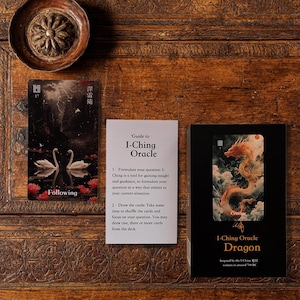 May include: An I-Ching Oracle card deck set featuring a black box with a dragon illustration, a white instructional guide card, and a rectangular card depicting two white swans on dark water with the text Following and the number 17.