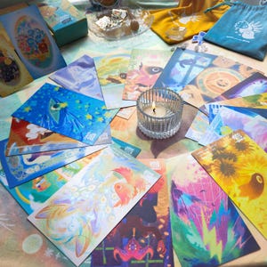 May include: A collection of illustrated tarot cards with colorful artwork, including images of celestial bodies, animals, and abstract designs. A lit candle in a glass holder sits among the cards, with small drawstring bags and a box in the background.
