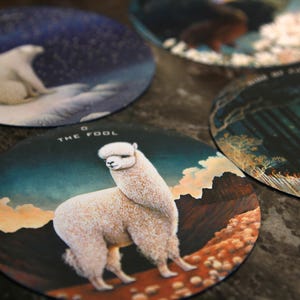 May include: A collection of round coasters featuring various illustrations. One coaster displays a white alpaca with the text "THE FOOL". Other coasters show a polar bear, a tiger, and other nature scenes.