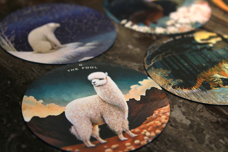 May include: A collection of round coasters featuring various illustrations. One coaster displays a white alpaca with the text "THE FOOL". Other coasters show a polar bear, a tiger, and other nature scenes.