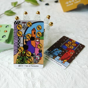 Puede incluir: Dos cartas de tarot, Diez de Pent&aacute;culos y Cinco de Pent&aacute;culos, con ilustraciones coloridas de gatos, perros y personas. La carta del Diez de Pent&aacute;culos presenta una familia de gatos y un perro de pie frente a una casa con una cerca y un jard&iacute;n. La carta del Cinco de Pent&aacute;culos presenta a tres personas caminando en la nieve, una de ellas con una capa roja con capucha.
