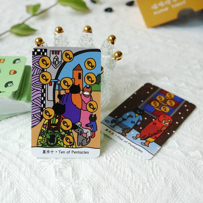 Puede incluir: Dos cartas de tarot, Diez de Pent&aacute;culos y Cinco de Pent&aacute;culos, con ilustraciones coloridas de gatos, perros y personas. La carta del Diez de Pent&aacute;culos presenta una familia de gatos y un perro de pie frente a una casa con una cerca y un jard&iacute;n. La carta del Cinco de Pent&aacute;culos presenta a tres personas caminando en la nieve, una de ellas con una capa roja con capucha.