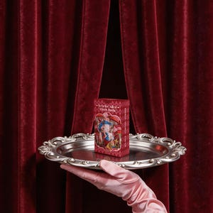 May include: A rectangular Scarlet Alice Tarot Deck box featuring a colorful illustration of a character in a red dress, resting on a round, ornate silver-toned metal tray with a decorative scalloped edge and a polished, reflective surface.