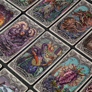 May include: A collection of rectangular playing cards featuring intricate, colorful illustrations of various mythical creatures and cosmic entities, each enclosed in a detailed, ornate, silver-toned border with specific names printed at the bottom.
