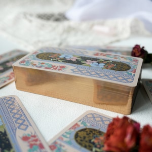 May include: A deck of tarot cards with a gold gilded edge and a colorful floral design on the back. The cards are stacked on a white surface.